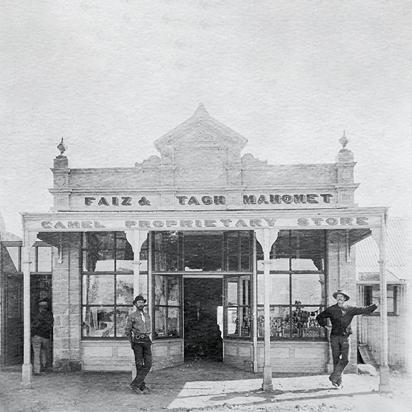 Picture of two men standing in front of the Faiz & Tagh Mahomet Camel Proprietary Store. They are leaning on posts either side of the entrance to the store looking at the camera, and are both wearing hats and work clothes. The one on the left has a beard and is wearing a short, wide tie. The one on the right has a moustache and is smiling broadly. The store