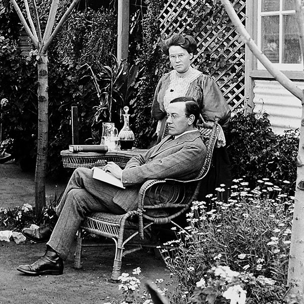 A picture of a besuited and bespectacled man, Dr Robert Mitchell, sitting, crossed-legged in a wicker chair, with an open book on his lap looking out to the left of the picture. He is in a flourishing garden in front of his home in Coolgardie. A bespectacled lady, in a high necked full-sleeved dress, is standing directly behind Dr Mitchell looking towards the camera. Photograph in possession of the author.