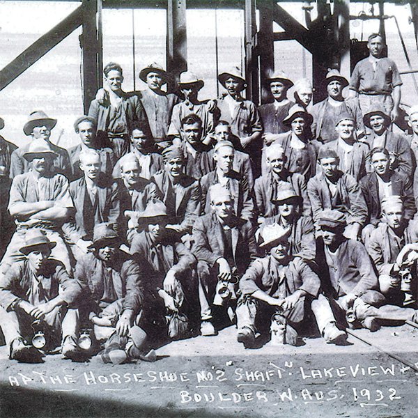 A picture showing a large group miners in work attire (in four rows—some sitting, some standing) at the Horseshoe No. 2 Shaft of the Lake View and Star Gold Mine, Boulder, 1932. Some men are holding their crib bags and carbide lamps. Photograph courtesy of Moya Sharp, OFH.