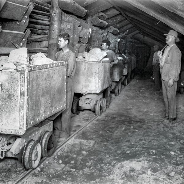 A picture showing men loading ore from stope chutes, on the left, into manually operated trucks in an underground mine circa 1920. The five bare-headed men, in work clothes, stand behind the rectangular, four-wheeled, trucks that have large lumps of rock protruding from them. To the right there is a man with a moustache and a broad-rimmed hat who appears to be overseeing the operations. Photograph courtesy of Moya Sharp, OFH.