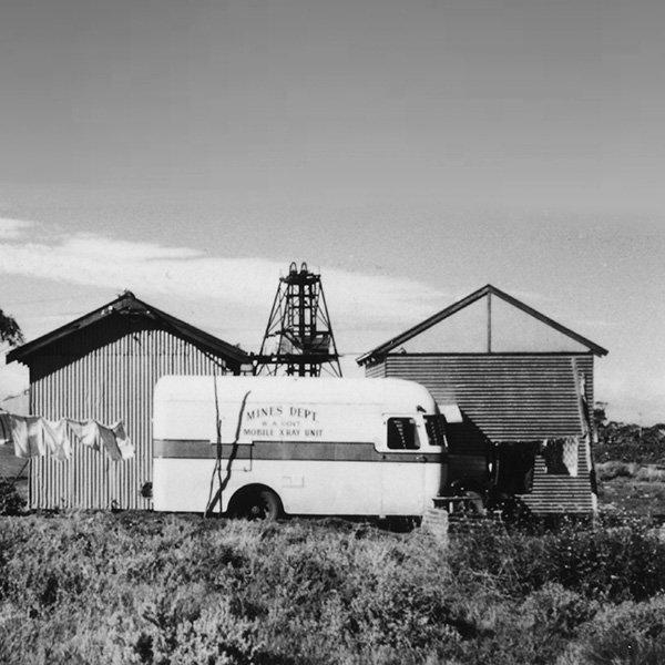 A picture showing the Mines Department mobile X-ray van at Calcion Gold Mine, Mt Ida. The van is in the centre of the picture, parked in front of two tin sheds with the mine