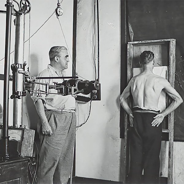 Photograph of a Gwalia miner, on the right-hand side of the picture, undergoing an X-ray examination. He is shirtless, with his back to the camera and hands on hips. On the left there is a middle aged man standing behind the X-ray equipment that consists of a horizontally mounted cylinder, approximately the width of the man