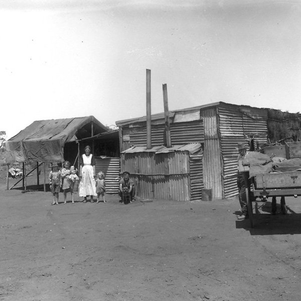 Picture showing a family in front of their home made of corrugated metal sheeting. To the left, there is a lady with four children and a dog, and on the right there