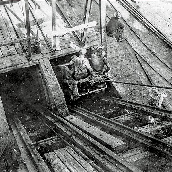Picture, taken from above, showing three mineworkers in a skip at the mouth of a timber built mine shaft. There are two entrances to the shaft, the skip is at the entrance to the one on the right, and is being propelled by a winch line along iron rails.