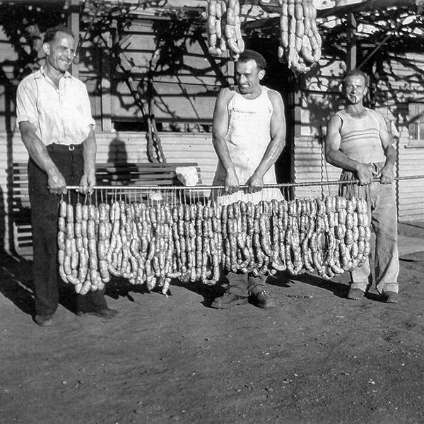 Picture showing three smiling men, Laurie Tognali, Varischetti and Mario Ponsanoli, holding, horizontally, a two metre long metal pole which has many strings hanging from it, each containing dozens of home-made ‘Italian’ sausages made from kangaroo, goat and donkey. They are pictured in front of a house in Gwalia circa 1940s.