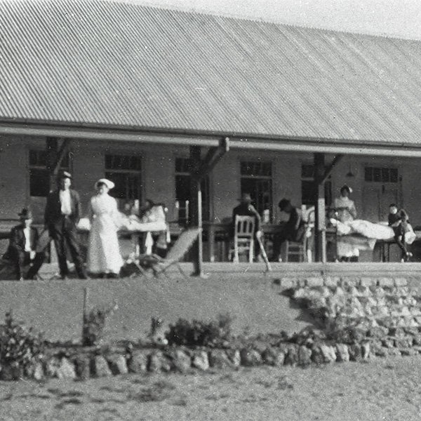 Picture showing patients and nurses on the veranda of the Wooroloo Sanatorium. The photograph has been taken some distance away from the building, with the grounds of the Sanatorium in the foreground. Some patients are standing, some are seated at a table and one is in a hospital bed, outside on the veranda.