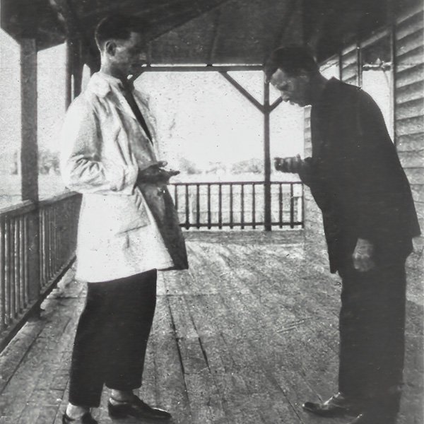 The picture of a patient undergoing a sputum test in an outdoor setting—on the veranda of a wooden building. The patient is a middle aged man in a dark jacket and trousers, who is sideways on to the camera on the right of the picture. He is leaning forward and his head is over an unseen object which he is holding in front of him in his right hand. He is faced by a younger, slightly taller man in a white lab coat, on the left of the picture. 