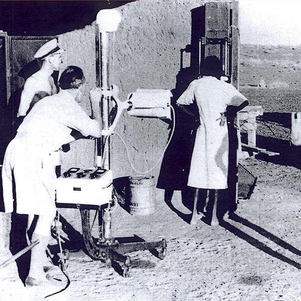 Picture depicting an X-ray being performed on a female member of a remote community. The very thin lady in a long white gown has her back to the camera and has her hands on her hips. She is facing part of the equipment which is against an outside wall, and there is very sparse vegetation in the landscape to the horizon. The machine, which has a central metal support, a control panel with large dials and knobs, and a rectangular unit for capturing the image, is being operated by a middle aged man in shirt sleeves and knee-length shorts who is bent over, lining up the equipment behind the woman. Photograph courtesy of Dr Alan King.