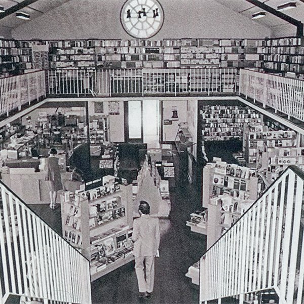 Picture taken from the top of a stairway looking down into the University of Western Australia Bookshop, 1975. There is a large open space on the lower level containing well-spaced bookstands with people browsing the books. The top level mezzanine floor has bookshelves along the lengths of the three visible walls.